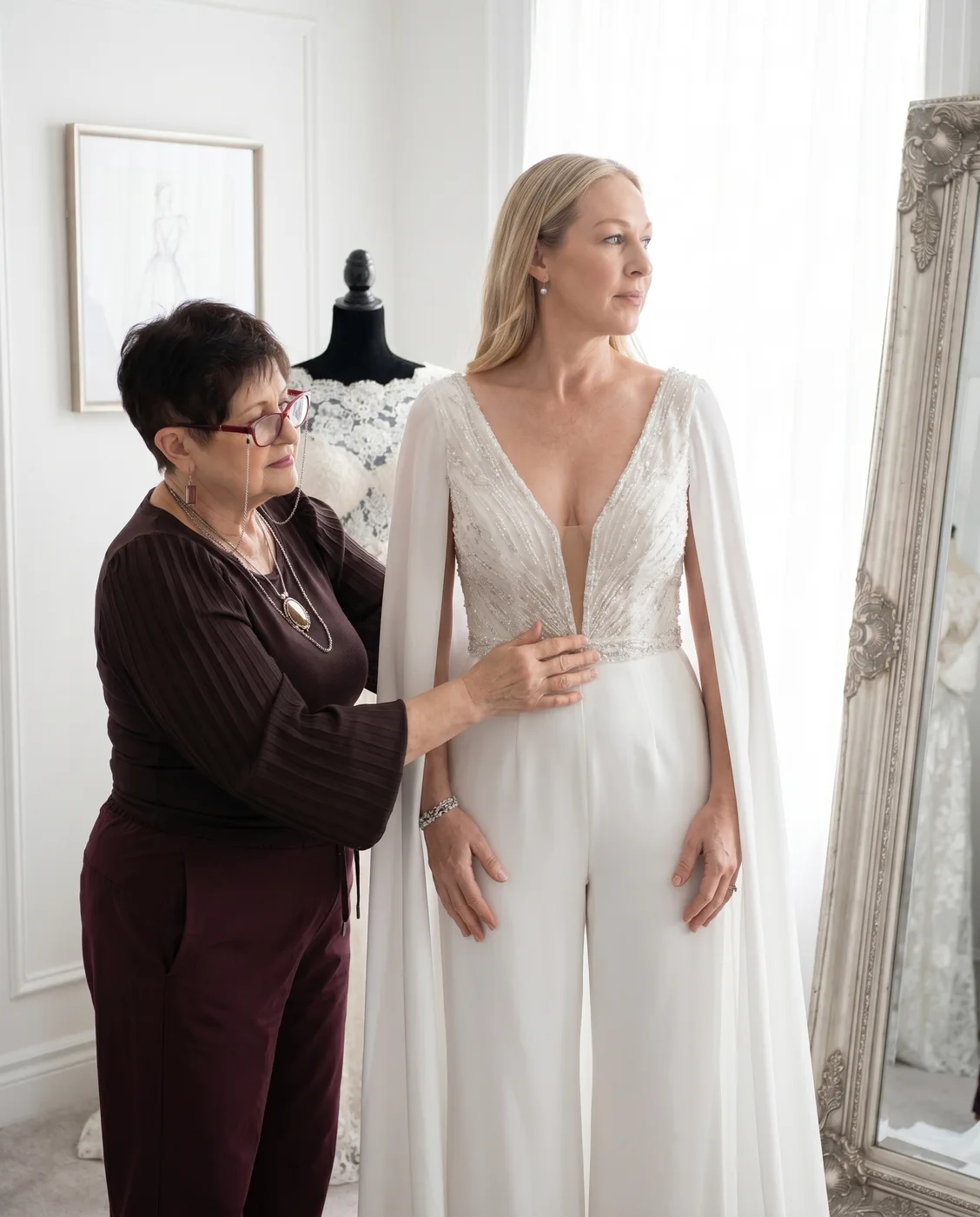 Designer Rita Kleiner fitting a client in an ivory beaded-cape bridal jumpsuit before the atelier mirror